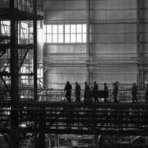 Workers wearing helmets walk along a metal walkway inside a large industrial factory.