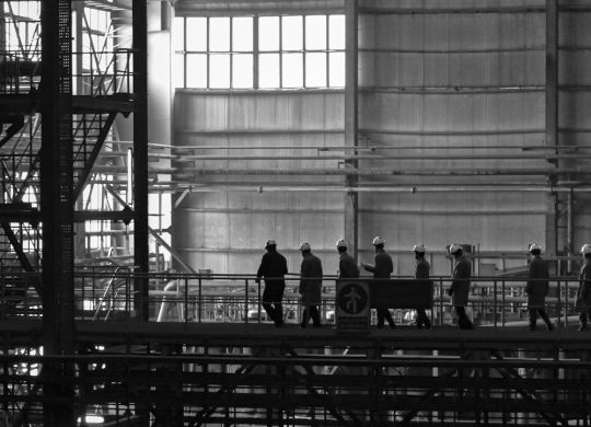 Workers wearing helmets walk along a metal walkway inside a large industrial factory.